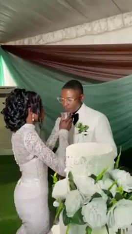 At a wedding reception, a bride and groom stand behind a three-tier white cake adorned with white roses. The bride, wearing a fitted white lace gown with long sleeves and her hair styled in an updo, holds a piece of cake to the groom's mouth. The groom, wearing a white suit with a black bowtie and glasses, appears to be emotional, wiping his eye as he takes a bite of the cake. The background features draped fabric in shades of brown and green, and an air conditioning unit is visible. The setting suggests an indoor wedding venue.