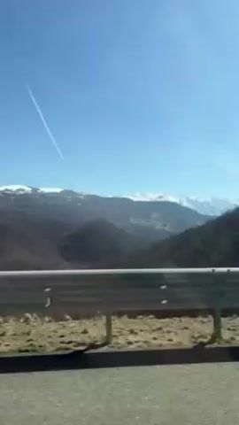 The video shows a view from a moving vehicle. The camera focuses on a metal guardrail, and beyond that, there are wooded hills and snow-capped mountains in the distance under a clear blue sky. A contrail is visible above. In the foreground, there is a traffic light and a tall metal structure with a ladder. An orange box-like structure is also visible near the guardrail. The sun is shining brightly.