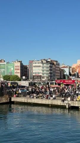 The video captures a daytime view of a bustling waterfront area with numerous people gathered on a pier or dock. The scene is set against a clear blue sky and features a variety of buildings in the background, ranging from low-rise to mid-rise structures. The buildings display a mix of architectural styles and colors, with some sporting billboards and advertisements. The pier is constructed of concrete and is lined with metal railings. People are seen sitting, standing, and walking around, suggesting a busy transit hub or public gathering spot. The water in the foreground is calm and reflects the daylight. Overall, the video portrays a lively urban waterfront environment on a clear, sunny day.