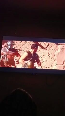 The video displays a screen showing a film. In the first frame, two men in Roman-era attire are in a desert-like environment. One man, wearing a helmet and armor, is facing away from the camera, while the other man, in a leather vest and kilt, is facing the camera, holding a weapon. The second frame shows the man in the leather vest, identified as an actor, holding a sword and looking to his left. The third frame shows him from the back, engaged in some action within a sandy or rocky area. The fourth frame shows the same actor from the front, holding a sword and looking forward, with a bright light or explosion in front of him. The film appears to be an action or historical drama.