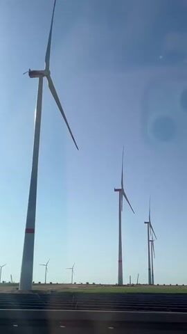 The video shows a row of wind turbines against a clear blue sky. The turbines are tall and white with red bands around their bases. The blades of the closest turbine are visible and appear to be spinning. The video is shot from a moving vehicle, as evidenced by the blurred foreground and the reflection of a phone in the sky. In the background, more wind turbines are visible in the distance, as well as fields of vegetation. The overall impression is one of a vast, open landscape with a wind farm dominating the scene.