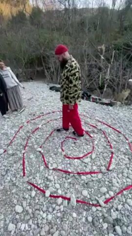 In an outdoor setting, a bearded man wearing a red beanie, a beige camouflage jacket, and red sweatpants is walking inside a large, spiraling shape. The spiral is created with red ribbon held in place by small rocks on a light gray, rocky ground. A few other individuals are standing at the side of the spiral. The background shows a dense area of shrubs and small trees. The lighting suggests either early morning or late afternoon, casting soft shadows on the scene.