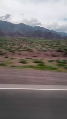 The video shows a blurred view from a moving vehicle. The scene is a wide landscape with rolling hills and mountains in the background under a cloudy sky. In the foreground, there is a paved road with a white line on the right side, indicating the edge of the lane. The road is dark gray. To the right of the road, there is a reddish-brown area with sparse green vegetation, appearing as blurred patches. Beyond this, there is a wider expanse of green vegetation, also blurred, leading up to the base of the mountains. The mountains themselves are a muted purple and brown, with their peaks softened by the distance and the cloudy atmosphere. The overall impression is one of motion and open, arid terrain. There are no people or specific objects visible.