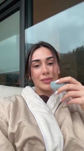 The video opens with a close-up shot of a young woman looking directly at the camera. She has dark hair pulled back loosely, with some strands framing her face. She is wearing a beige or light brown bathrobe with a white collar and lapels. The letters "LAN" are visible on the right side of her bathrobe. She holds a small, light blue-gray cup to her lips, seemingly taking a sip. Her fingernails are painted a light, natural pink color. Behind her, large glass windows or doors are visible, reflecting parts of the indoor space. A white cushioned chair or sofa is next to her.

As the video progresses, the camera pans to the left, revealing more of the indoor setting and then shifting focus to the view outside the large glass doors. A spacious wooden deck or balcony is visible immediately beyond the doors. The deck appears wet, suggesting recent rain. On the deck, near a low stone wall, are two dark-colored decorative lanterns with glass panels.

The outdoor scene is dominated by a misty, mountainous landscape. Rolling hills covered with various trees, some with bare branches and others with budding green foliage, stretch into the distance. A soft, hazy fog or cloud cover hangs low over the mountains, creating a serene and somewhat muted atmosphere. The overall impression is one of quiet relaxation in a natural, possibly resort-like, setting.