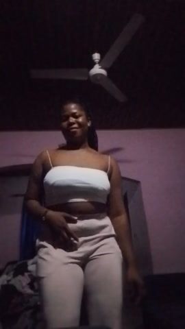 A woman stands in front of a pink wall. She wears a white strapless top and light-colored pants. Her hair is dark and styled in braids. She is facing the camera, with her left hand on her hip and her right hand resting on her stomach. A ceiling fan is visible in the background to the right of the woman. The lighting in the room is dim.