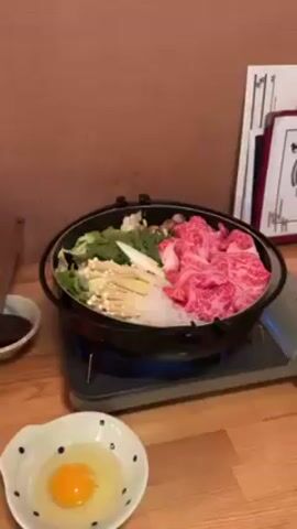 A tabletop sukiyaki setup is shown.  A burner is under a sukiyaki pot containing vegetables, mushrooms, rice noodles, and marbled raw beef. A bowl containing an egg is in the lower left corner of the frame, and a small bowl with a brown sauce and a long handled spoon is on the upper left. The background is a tan wall with framed text documents on it. No people are visible.