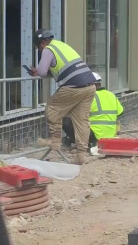 The video shows two construction workers on a site. The worker in the foreground is wearing a hard hat, a light purple long-sleeved shirt, a bright yellow safety vest with reflective stripes, tan work pants, and tan work boots. They are holding a dark-colored smartphone in their left hand and appear to be looking at it. They are leaning on a metal barrier. Behind them, a second worker is wearing a white hard hat and a bright yellow safety vest, and is kneeling on the ground. The environment appears to be a construction site with gravel and red plastic barriers visible in the foreground. The background shows a building under construction with windows and brickwork. A reflection in the window shows another worker in an orange high-visibility vest. The lighting suggests it is daytime.
