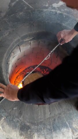 The camera provides an overhead view into a tandoor oven filled with hot coals and flames. At the bottom of the oven is a metal dish filled with food. A metal rod with a loop is inside of the tandoor. The oven itself is made of brick and is located inside of a room with tiled flooring. A cloth lies in the background of the shot.