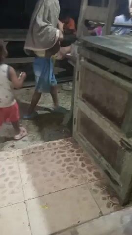 A video shows a young child in a white shirt with a pattern and pink shorts walking away from the camera. Next to the child is an older child wearing a light-colored headscarf, blue shorts, and a white shirt, holding something white to their face. The older child is standing next to a wooden structure that appears to be a table with benches. The ground is tiled and wet in some areas. The scene is dimly lit, suggesting it is nighttime or indoors with low lighting.