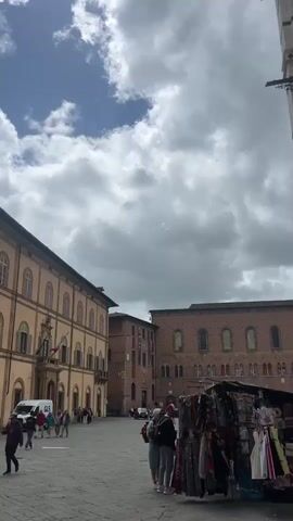 The video shows the exterior of a large historic building in Italy. The building is made of brick and stone, and has a number of arched windows and doorways. There are people walking around in front of the building. The sky is partly cloudy. There is a white van with the letters "GLS" written on the side, parked on the left of the scene. The video pans to show the columns of a portion of the building that is made of white stone and features alternating black stone details.