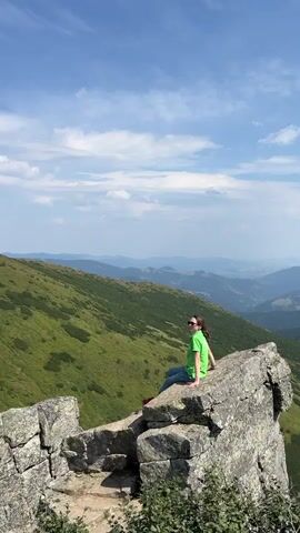The video shows a person sitting on a large rock formation on a mountainside. They are looking out at a vast landscape of rolling green hills and mountains under a blue sky with scattered clouds. The camera pans across the landscape, showing more of the mountain range and the distant horizon. The person is wearing a green shirt and blue jeans. The rock formation is rough and uneven, with some vegetation growing around its base. The overall scene is one of natural beauty and tranquility.