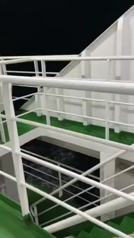 The video shows a nighttime view from the deck of a ship. The camera is pointed down through a white railing towards the dark water below, revealing the ship's wake.  The deck is green and yellow, and the railings and walls are painted white.  There are steps visible leading down, suggesting the presence of multiple levels on the ship.