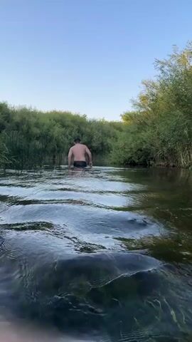 A person wearing black swim trunks walks away from the camera into a shallow, clear river. The person is facing away from the viewer, and their back is to the camera. The river is surrounded by dense green foliage on both sides, and the sky is clear blue. The water is calm with small ripples on the surface, and aquatic plants are visible at the bottom. The camera is positioned low to the water, giving a unique perspective of the scene. The lighting suggests it is late afternoon or early evening, with the sun casting a warm glow on the environment.