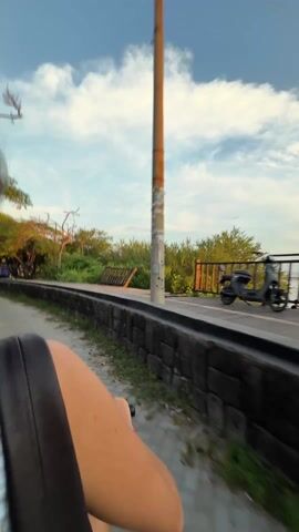 A person in a blue and white checkered shirt and a black bag is a passenger on a scooter traveling down a paved road. The scenery includes trees, a wooden boardwalk next to a body of water, and a woman driving a red and white scooter. In a close-up view, the passenger films another woman riding a scooter ahead of them, dressed in a white dress.  Other people are seen in the background. The light suggests it is late afternoon or early evening.