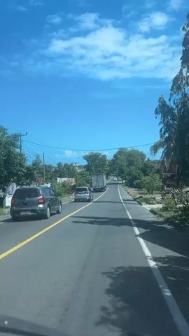 The video shows a perspective from inside a vehicle traveling on a road. The road is paved with a yellow line marking the center and a white line on the right edge. There are several other vehicles on the road, including cars and trucks. The surroundings are lush with green trees and some buildings are visible in the distance. The sky is bright blue with scattered white clouds. The overall impression is of a sunny day in a rural or semi-rural area.