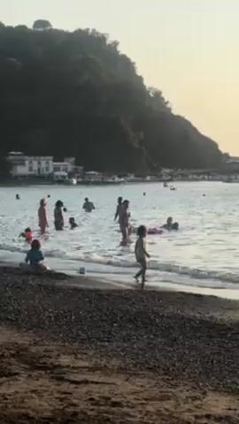 The scene captures a beach with a dark, pebbly shoreline where several people, including adults and children, are enjoying the water. Some individuals are standing and wading in the shallow water, while others appear to be swimming. One child is walking along the edge of the water, and another is playing near the shoreline with a bucket. In the background, there is a hill covered with trees and greenery. A building with a white exterior and multiple windows is visible in the distance, situated near a harbor or dock area where boats are parked. The lighting suggests it might be late afternoon or early evening, as the sun casts a soft glow over the scene.