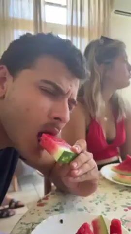 A young man with dark curly hair and a slight mustache is seated at a table indoors, intently eating a slice of watermelon. He is wearing a dark shirt and his face shows concentration as he chews. To his right, a blonde woman in a pink top sits facing away from the camera, seemingly observing something else. The table is covered with a floral patterned tablecloth, and several more slices of watermelon are visible on white plates. The room is well-lit, with sheer curtains hanging over a window in the background.