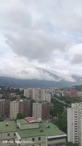 The video is a vertical, high-angle shot of a city skyline with mountains in the background. The sky is overcast with thick, grey clouds. In the foreground, several multi-story apartment buildings are visible, some with green roofs. Beyond the apartment buildings, more city buildings stretch into the distance, and a highway overpass can be seen. The mountains are partially obscured by clouds, with only the lower slopes visible. The overall impression is of a cloudy day in a densely populated urban area with a mountainous backdrop. The video has a watermark at the bottom left corner that reads "infinix HOT 50Pro".