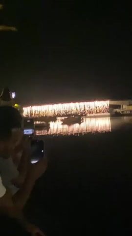 A night-time video shows a body of dark water reflecting the lights of a long, illuminated building or bridge in the distance. Several boats are visible on the water in front of the structure. In the foreground, the hands of two people are visible holding up cell phones, presumably to record the scene. The lighting is low, and the overall atmosphere is dark.