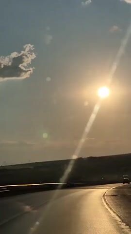 This is a vertical video clip filmed from a moving vehicle, likely a car, on a sunny day. The viewpoint is from the front passenger seat, looking out through the windshield. The primary subjects are the sky, the sun, and a highway.

The scene is set on a highway under a bright, clear blue sky with a few fluffy white clouds. The sun is prominently featured, positioned high in the sky and casting a strong glare, creating lens flare effects and streaks of light across the frame. The sun's brilliance suggests it is either late morning or mid-afternoon. The highway curves gently to the right, its asphalt surface reflecting the golden light of the sun. In the distance, the landscape appears flat and somewhat barren, with hints of vegetation and what might be a distant town or structures. A few vehicles are visible on the highway ahead, their shapes indistinct due to the distance and the glare.

The overall mood is serene and perhaps contemplative, with the vastness of the sky and the open road evoking a sense of journey and freedom. The bright sunlight and clear skies indicate fair weather. There is no visible text in the video clip. The mention of Gorno Kamartsi, Bulgaria, places the setting geographically.