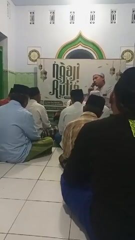 The video shows a group of men sitting on the floor, facing a man who is speaking. The setting appears to be a room with white walls and a tiled floor. There is a banner in the background with text that reads "Ngaji Rutin". The men are dressed in traditional Islamic attire, including peci hats and sarongs. Some of the men are looking at the speaker, while others are looking down. The overall atmosphere seems to be one of a religious gathering or a study session. The video is shot from a slightly elevated angle, showing the backs of most of the men in the foreground. The lighting is bright and even, indicating an indoor environment. There is a window visible on the left side of the frame, with green bars.