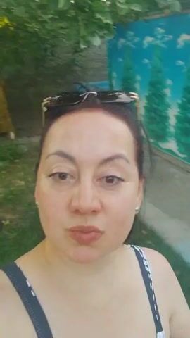 A woman in a black and white swimsuit with sunglasses on her head films herself in a selfie-style video. She is outdoors, with a mural of green trees and a blue sky visible in the background. To her right, a person in a patterned outfit walks by, and further in the background, a swimming pool is visible. The woman moves her arms and the camera pans around her.