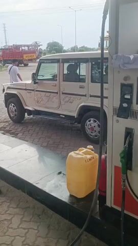 The video shows a light-colored Mahindra Bolero parked at a diesel pump. A man wearing a white shirt and blue jeans stands near the driver's side door of the Bolero, his hands on his hips. The fuel pump displays "DIESEL" and has two nozzles. One nozzle is attached to a hose that is resting on a yellow jerrycan placed in front of the pump. The pump screen shows a transaction amount of 510.00 with liters displayed as 4.82. A red fire extinguisher is visible next to the pump. In the background, a yellow truck with a red body is parked. The environment appears to be an outdoor fuel station with a paved ground.