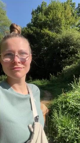 A young woman with blonde hair pulled into a bun and wearing glasses and a light teal t-shirt stands in an outdoor setting with greenery and trees surrounding her. She is looking at the camera and smiling slightly. A dog is partially visible to her right. The woman is carrying a light-colored bag over her shoulder. The video appears to be a selfie taken while walking.