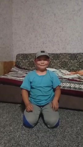 A young boy wearing a blue t-shirt, gray pants with blue accents, and a grey baseball cap is kneeling on a grey carpet. He is looking directly at the camera with a slight smile. Behind him is a couch with a patterned upholstery and a patterned bedspread. The wall behind the couch has a textured wallpaper. The scene appears to be indoors, likely in a living room or bedroom. The lighting suggests it is daytime. No visible text is present. The boy's expression and posture suggest a casual and possibly playful mood. The location context is Chon-Kara, Kyrgyzstan.