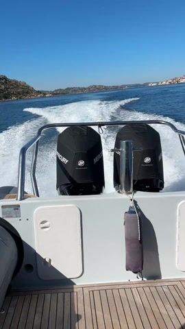 The video shows a view from the stern of a boat moving across a body of water on a clear, sunny day. Two Mercury outboard motors, covered in black protective covers, are prominently displayed at the back of the boat. The boat's wake is visible behind it, creating a white foamy trail on the blue water. The boat has a white and gray exterior, with a wooden deck. In the distance, there are land masses with rocky terrain, and some buildings can be seen along the coast.