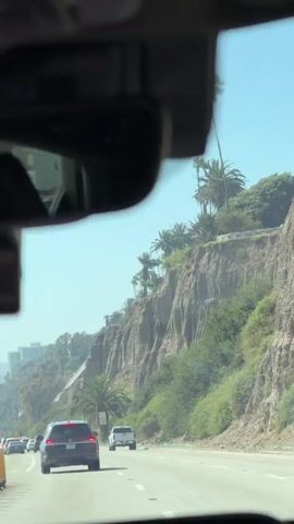 This media file captures a scenic drive along a coastal highway, likely in Santa Monica, California, given the presence of palm trees, steep cliffs, and ocean-adjacent scenery. The perspective is from inside a vehicle, looking out through the windshield and rearview mirror.

The primary subjects are the vehicles on the road, including a dark SUV in the foreground, a white pickup truck, and several other cars ahead. The scene is dominated by a dramatic cliff face covered in lush green vegetation and dotted with tall palm trees, suggesting a warm, sunny day with clear skies. The road appears to be a multi-lane highway, with traffic moving steadily.

Notable details include a speed limit sign indicating "45," visible on the right side of the road, and various trees and buildings in the distance, including what appear to be apartment buildings or hotels, hinting at an urban or suburban environment bordering the natural landscape. The bright sunlight and clear blue sky indicate it is daytime, likely late morning or early afternoon.

There is no discernible emotion conveyed in the media, as it is purely observational. The activity is simply driving, with the camera capturing the view from the passenger or driver's seat. The overall impression is one of a pleasant journey through a visually striking environment.