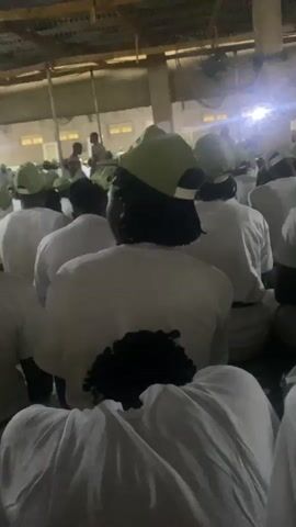 The video shows a crowded indoor space, possibly a hall or gymnasium, with a corrugated metal roof supported by wooden beams and metal poles. The lighting appears artificial and somewhat dim, with bright spotlights casting illumination. The majority of the people visible are seated on the floor, facing away from the camera. They are all dressed in white t-shirts and green baseball caps. The camera is positioned behind a few individuals, focusing on their backs and heads. Some people appear to be interacting, with one person in the foreground turning their head to speak to someone to their side. The overall impression is one of an organized gathering or event, possibly a training session or orientation, given the uniform attire. The environment seems somewhat utilitarian, with exposed infrastructure elements.