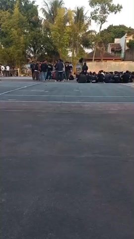 The video shows an outdoor scene, possibly a sports court, with several groups of people. In the foreground, the court surface is visible, appearing to be asphalt or concrete. In the middle ground, a group of young men, some wearing black clothing, are gathered and appear to be talking or listening to someone. A net, possibly for a sport like volleyball or badminton, is set up across the court. Further in the background, more people are visible, including some standing and others sitting on the ground. Trees and buildings are present in the background, suggesting an urban or suburban environment. The lighting suggests it is daytime, possibly late afternoon or early evening. One person in the foreground is walking across the court.