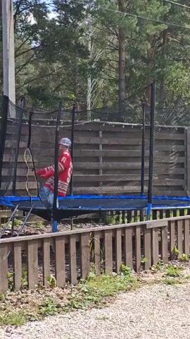 A person in a red hockey jersey with the number 91 on the back and a white baseball cap is jumping on a trampoline. The trampoline is blue and black and has a safety net. The person is wearing jeans and black shoes. The trampoline is in a backyard with a wooden fence and trees in the background. The person is jumping up and down and appears to be enjoying themselves.
