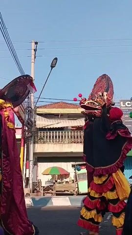 A street scene with people watching a parade. In the foreground, a child wearing a black t-shirt with blue lightning patterns and the word "Madura" on the back is visible. Behind the child, there are performers in elaborate costumes, likely part of a traditional dance or procession, characterized by vibrant colors, masks, and ornate headdresses. One performer is dressed in black with red, yellow, and white ruffled accents. Other individuals, some appearing to be spectators and possibly performers or organizers, are scattered throughout the scene. A man in a gray and teal shirt and a blue cap holds a plastic bag filled with items. A green food cart is parked on the side of the street, and buildings line the background. Utility poles and wires are also present. The overall atmosphere suggests a public event or celebration.