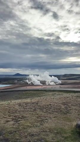 The image presents a wide-angle landscape view of a geothermal area in Reykjahlíð, Iceland. In the foreground, a gently sloping hillside covered with dry, sparse brown and green vegetation leads down to a paved asphalt road that curves across the midground.

Beyond the road, a broad, flat plain is dominated by large, white plumes of steam rising vigorously from the ground, indicative of significant geothermal activity. These plumes likely originate from a geothermal power plant or natural vents, with industrial-looking structures, including a small red building, partially obscured by the steam. To the left of the main steam clouds, a distinct, light blue body of water, possibly a geothermal lagoon or cooling pond, is visible.

The background features a large, dark body of water, likely a lake, with distant hills or mountains lining the horizon under a vast, cloudy sky. The sky is heavily overcast with diffused light, though a brighter patch is visible on the right side of the frame, suggesting a partly cloudy day.

No people or discernible text are present in the scene. The overall impression is one of raw natural power within a barren yet striking landscape, characteristic of Iceland's volcanic regions.