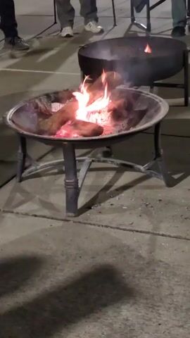 The video shows a metal fire pit on a concrete surface. The fire pit contains burning logs and small flames. Behind it, another fire pit, also metal, has visible smoke emanating from it. In the background, parts of several people are visible from the waist down. They are wearing various types of shoes and pants. The lighting suggests the scene is at dusk or night.