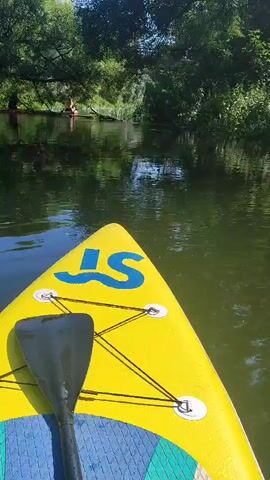 The video shows a first-person perspective of someone on a yellow paddleboard with a blue logo. The paddleboard is on a calm body of water, likely a river or lake, with lush green trees and vegetation lining the banks. In the distance, other people can be seen on paddleboards or small boats. The water is dark and reflects the surrounding greenery. The overall atmosphere is peaceful and suggests a recreational activity in nature.