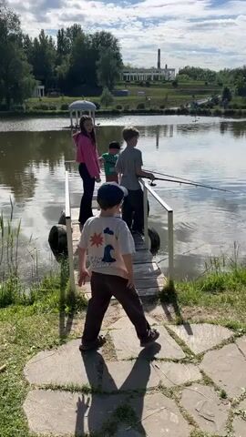 Four children are fishing on a small wooden dock, with a fifth child walking away from the dock. The dock extends over a lake, and a small white gazebo is visible on the water to the left. In the distance across the lake, there's a large white building with a tall smokestack. The sky is partly cloudy with sunlight breaking through.