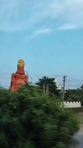 This video shows a large, orange statue of a Hindu deity, possibly Hanuman, as viewed from a moving vehicle. The statue is seen from its back and side, with a golden crown or headdress. The statue is situated amidst lush green foliage and trees, with power lines visible in the background against a cloudy sky. The camera appears to be panning or moving, causing a slight blur in the foreground vegetation, suggesting the shot was taken from a car or similar mode of transport. In the later frames, a white building and a red fence can be seen in the distance behind the statue and trees. The overall scene is outdoors, under an overcast sky, with a focus on the prominent religious monument.