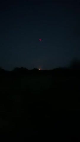 The video captures a dark, nighttime scene. In the upper portion of the frame, two red lights are visible, one directly above the other. Below these lights and closer to the horizon, there's a faint green glow. The lower half of the video is almost entirely black, with only a dark silhouette of the landscape visible. Towards the end of the video, a distant orange glow appears on the horizon, possibly indicating a sunrise or a light source behind the trees.