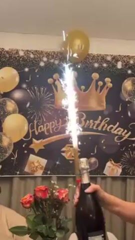 A video shows a person holding a large bottle of champagne with a lit sparkler fountain erupting from the top, against a "Happy Birthday" backdrop with gold and black decorations. The person is standing behind a table adorned with a vase of red and orange roses. Other people are visible in the video, but are out of focus. The scene suggests a birthday celebration.