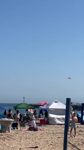 The video captures a beach scene under a clear blue sky. In the foreground, a beach volleyball net is set up, with several people visible on both sides of it. Some individuals are playing or preparing to play volleyball, while others are watching from the sand. Beach tents and umbrellas are scattered across the sand, providing shade for beachgoers who are relaxing on chairs or the sand. In the background, the ocean is visible, with waves and a boat making its way across the water. The overall atmosphere appears to be one of a lively and enjoyable day at the beach.