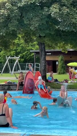 A crowded outdoor swimming pool area is depicted on a sunny day. In the foreground, several children and adults are in the bright blue water. A young child with blonde hair is in the center, looking towards the camera, with water splashing around them. To the left, a woman with blonde hair and a black swimsuit is seated on the pool edge, facing away from the camera. Further left, another child is in the water, with their arms outstretched. A red waterslide is a prominent feature in the middle of the scene, with a child in a blue hat sliding down it. Another child is at the bottom of the slide, in the water. To the right of the slide, several people are relaxing on the grass. A man with dark hair and a cap is sitting on a stone wall on the far left, looking towards the pool. Behind the slide and people in the pool, a large, leafy green tree provides shade. A wooden building with a dark roof is visible in the background, with a swing set to its left. Several other people are visible in the background, some sitting on the grass and others standing near the building. The overall atmosphere appears to be one of recreation and enjoyment.