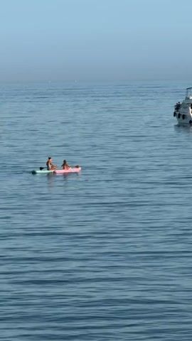 A white yacht is anchored in calm blue water under a clear sky. To the left of the yacht, two people are on paddleboards. One person is sitting on a pink paddleboard, and the other is sitting on a green paddleboard. They are facing each other and appear to be interacting. Another person is visible on a separate paddleboard behind them. Further to the right, a white sailboat is visible in the distance. The water is slightly rippled.