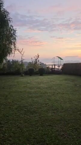 The video shows a sunrise over a body of water. The sky is filled with soft pink and orange clouds, and a gentle mist hovers over the water. In the foreground, a lush green lawn leads to a small dock with a covered seating area. Trees and bushes frame the scene, adding depth and texture. The overall atmosphere is peaceful and serene.