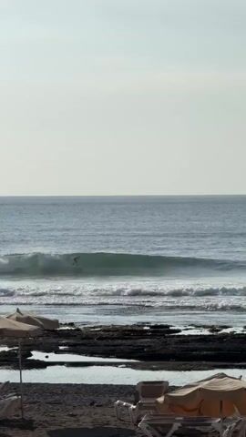 The video shows a beach scene with a surfer riding a wave in the ocean. The sky is overcast, and the sun is casting a soft light. In the foreground, there are beach chairs and umbrellas, suggesting a recreational area. The water is a calm blue, with moderate waves breaking near the shore. The beach itself appears to be rocky, with patches of sand. There are a few people visible on the beach, some standing and others possibly sitting or lying down. The surfer is the main subject of action, actively participating in the sport. The overall atmosphere is peaceful and relaxed, typical of a beach environment.