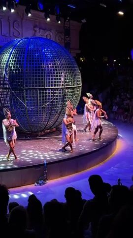 This video shows a performance on a stage with a large metal sphere behind several performers. The performers, appearing to be female, are dressed in light-colored, ornate costumes with headdresses and are performing synchronized dance movements on a circular stage. The stage is lit with blue and purple lights, and a large audience is visible in the background, silhouetted against the stage lights. The metal sphere is a cage-like structure. A motorcycle is partially visible next to the sphere. The text "LIS" is visible on a structure behind the stage.