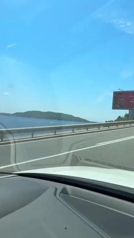 The video appears to be shot from inside a car, looking out the windshield on a sunny day. The car is on a highway, with the ocean visible to the left and a green, hilly landscape to the right. There is a billboard on the right side of the road. The sky is a clear blue with some wispy clouds. Traffic is visible on the road, including another vehicle, a white van. The video shows a scenic coastal drive.