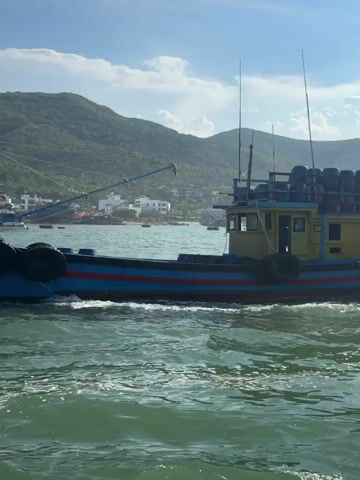 This media file captures a vibrant scene on the water, likely in Nam Nha Trang Ward, Vietnam. The primary subject is a brightly colored fishing boat, predominantly blue with red and yellow accents. It appears to be in motion, creating a wake in the choppy, green water.

The boat is loaded with what seem to be blue barrels or containers stacked high on its deck. There are also several tires attached to its sides, serving as fenders. Two tall antennas or fishing rods extend from the top of the boat. In the background, a larger, more modern white passenger boat is visible, along with other structures along the coastline. Beyond the immediate waterfront, lush, green, mountainous terrain rises under a bright, partly cloudy sky, suggesting fair weather and a daytime setting.

While no people are clearly visible on the fishing boat, the presence of fishing equipment and the setting strongly imply it is engaged in or returning from fishing activities. The overall mood is one of active coastal life, with a sense of industry and the beauty of a tropical or subtropical environment. No visible text is present in the frame.